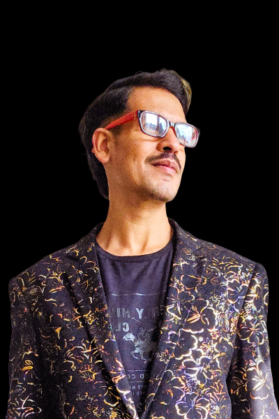 Portrait of a designer in red-framed glasses and a black jacket with a gold floral pattern, looking up and to the side with a slight smile.